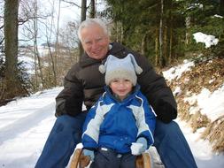 Smiling grandpa, with the cute, child, sliding on the toboggan, among the beautiful, snowy forest, in the winter