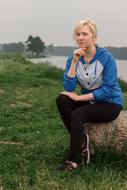 Blonde girl, sitting on the rock among the green grass, on the shore under cloudy sky