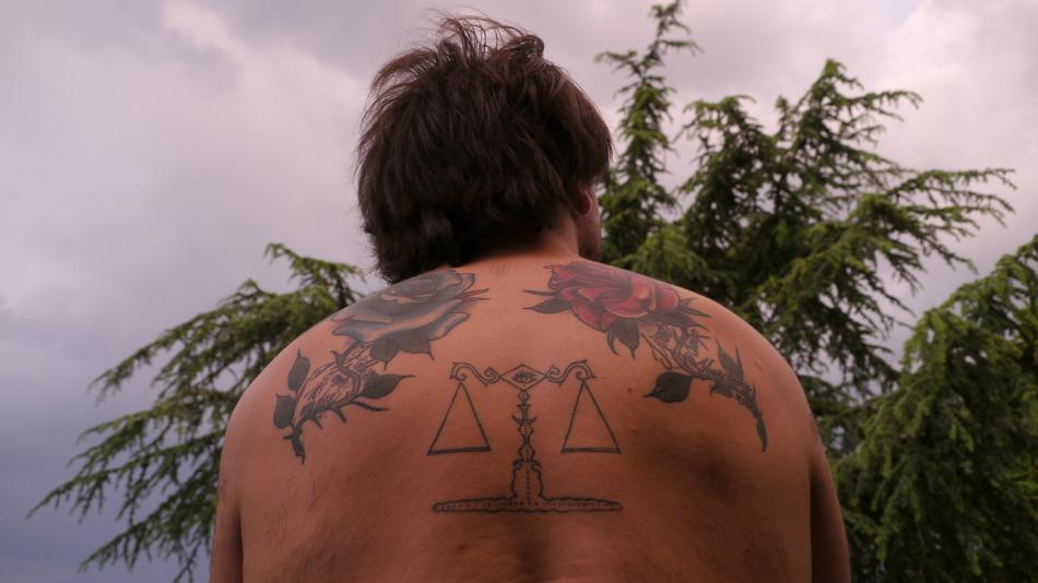 Back view of the man with tattoos, near the green tree, under the beautiful and colorful sky with clouds