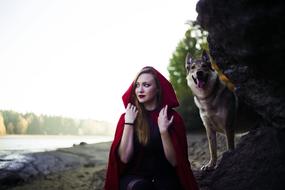 Girl in Little Red Riding Hood costume, posing with the dog, on the shore with plants