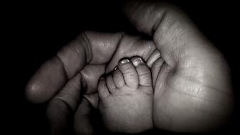 Black and white photo with close-up of the child foot in the adult's hand, at dark background