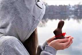 Bunny girl in hood, holding beautiful chocolate Easter bunny on the hands, near the water