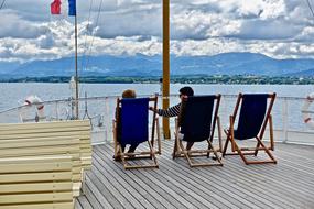 People sitting on the deck chairs on the yacht with flag of France, at background with the coast with mountains