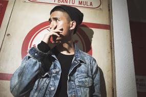 a guy in a denim jacket smokes near the wall of a building