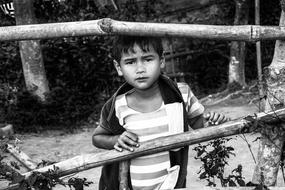 Black and white portrait of the kid boy standing behind the fence, among the plants